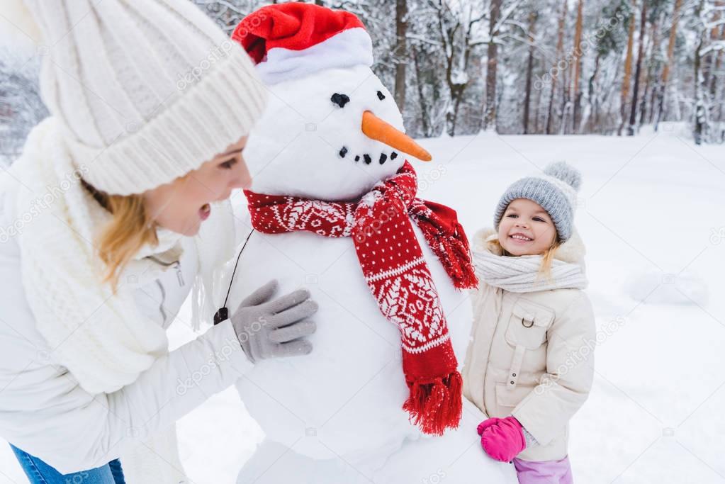 happy mother and daughter playing with snowman in winter park