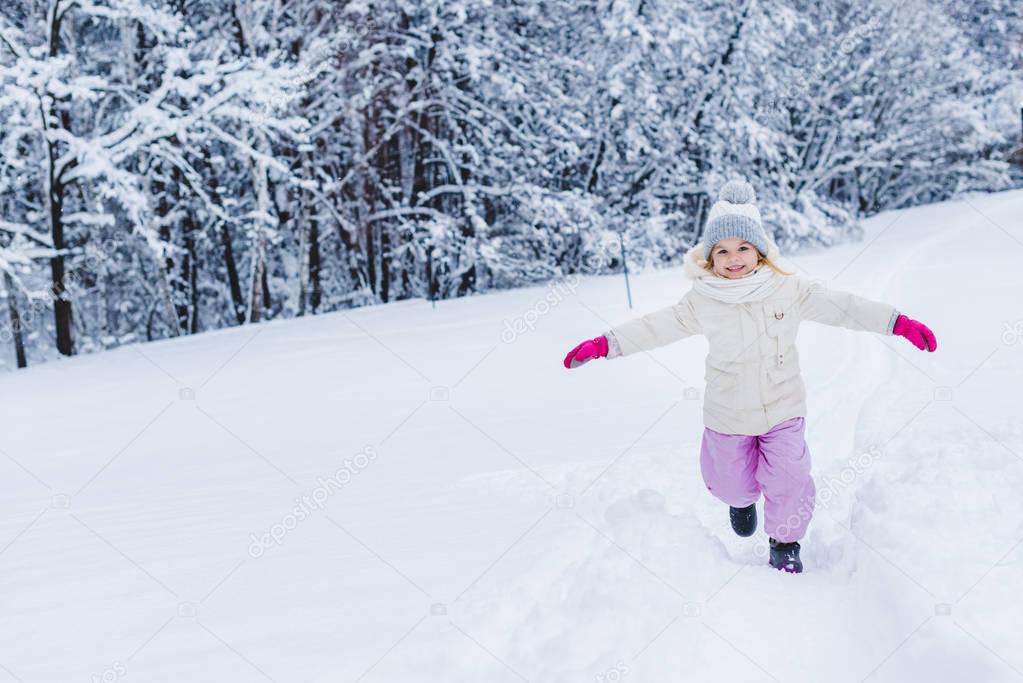 adorable happy child with open arms in mittens running in snow and smiling at camera in winter park   
