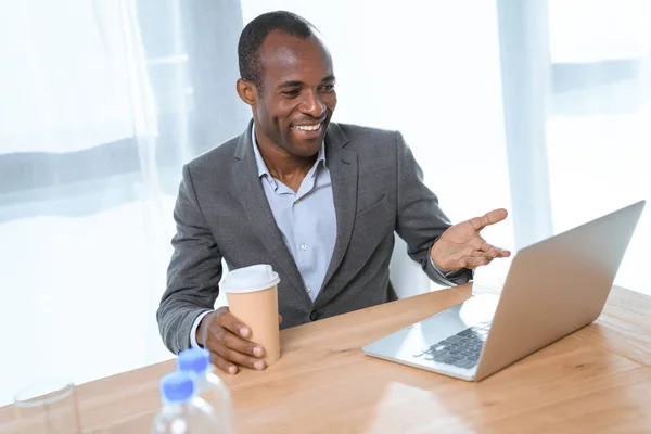 テーブルの上のノート パソコンを見ながら笑みを浮かべてコーヒー カップでアフリカ人の笑顔  — 無料ストックフォト