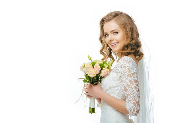 side view of smiling bride in wedding dress and veil with bouquet of flowers isolated on white