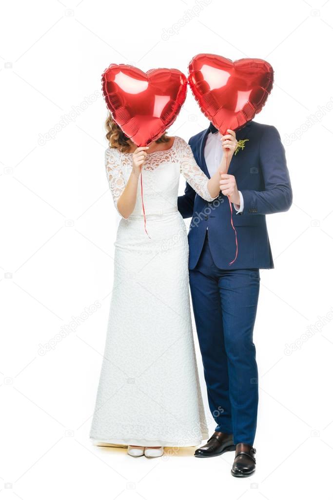 wedding couple covering faces with red heart shaped balloons isolated on white 