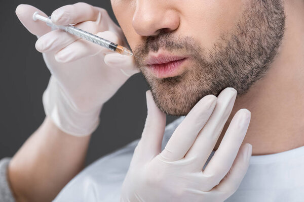 cropped view of hands in medical gloves making beauty injection for man, isolated on grey
