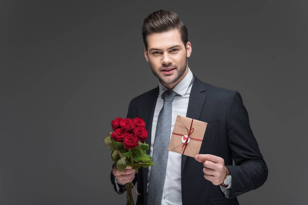 handsome man in suit holding red roses and envelope for valentines day, isolated on grey