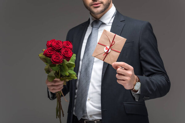 cropped view of man in suit holding red roses and envelope for valentines day, isolated on grey