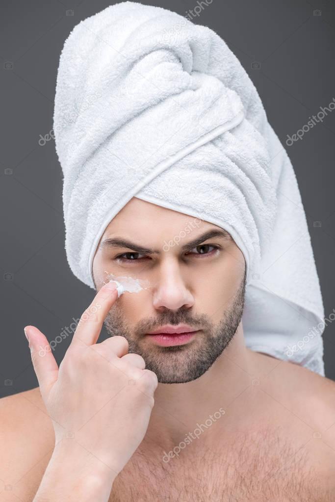 man in towel applying cosmetic cream on face, isolated on grey, skin care concept