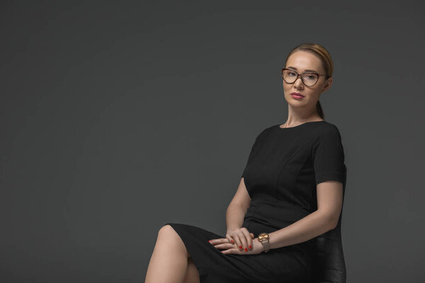 beautiful kazakh woman in eyeglasses sitting on chair and looking at camera isolated on grey