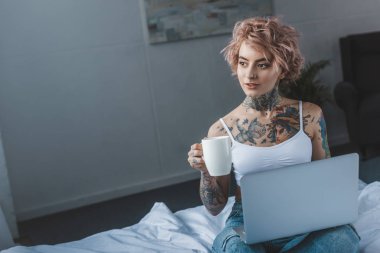 güzel dövmeli kız yatakta dizüstü kullanarak kahve ile 