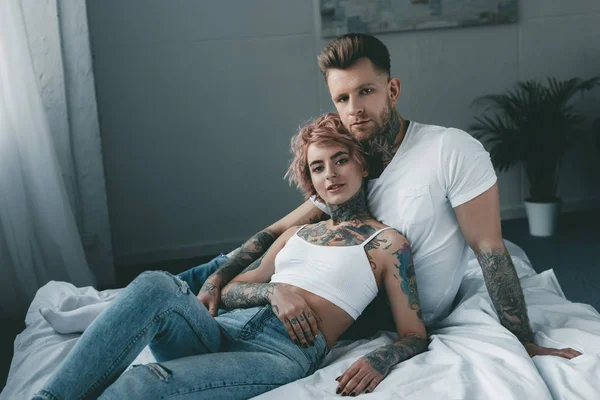 tattooed couple looking at camera and relaxing on bed