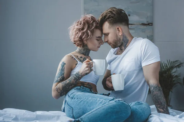 tender tattooed couple with coffee touching foreheads in bedroom at morning