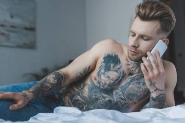 yatakta yatarken Smartphone'da konuşan yakışıklı gömleksiz dövmeli adam 