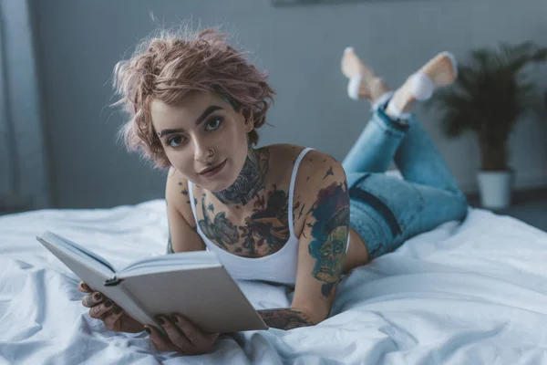 Стильная Татуированная Девушка Читает Книгу Кровати Смотрит Камеру — Бесплатное стоковое фото
