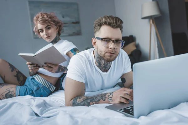 Tatuado Namorada Leitura Livro Enquanto Namorado Usando Laptop Quarto — Fotografia de Stock