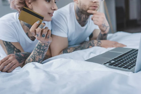 young tattooed couple shopping online with credit card and laptop in bed