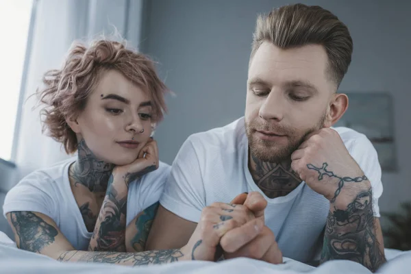 young tattooed couple lying on bed and holding hands