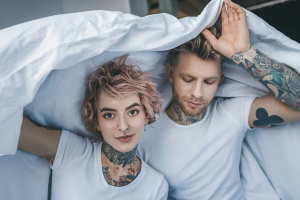 young tattooed couple under white blanket in bedroom