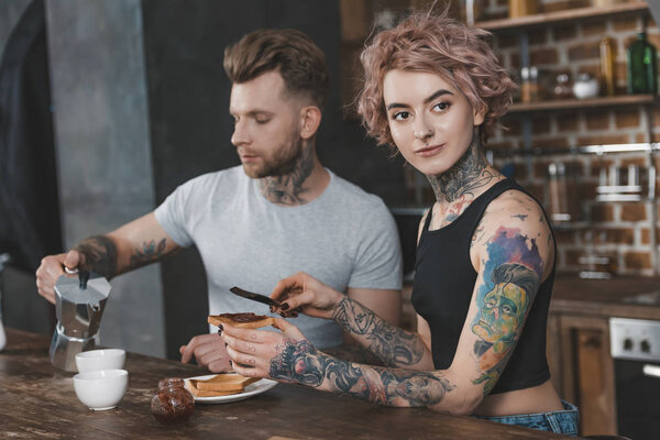young tattooed couple eating toasts and drinking coffee for breakfast 
