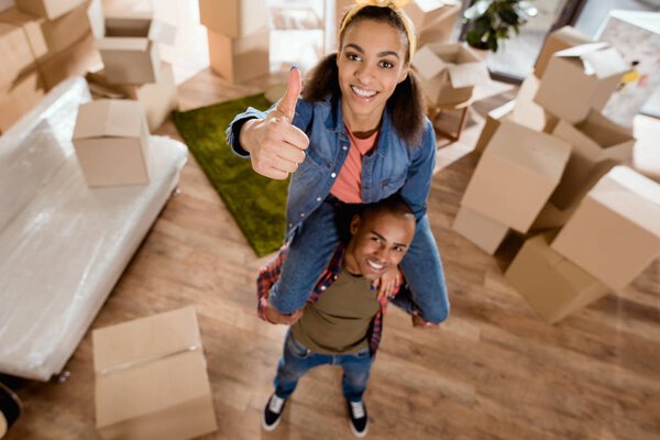 smiling african american girl sitting on shoulders of her boyfriend and showing thumb up in new home