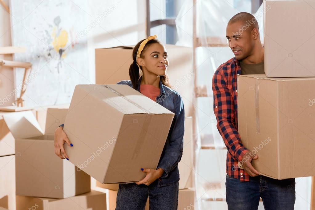 african american couple holding cardboard boxes and moving to new apartment