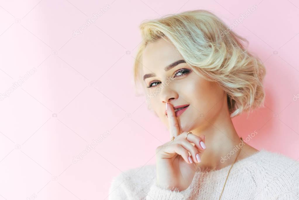 beautiful blonde girl gesturing for silence and looking at camera isolated on pink