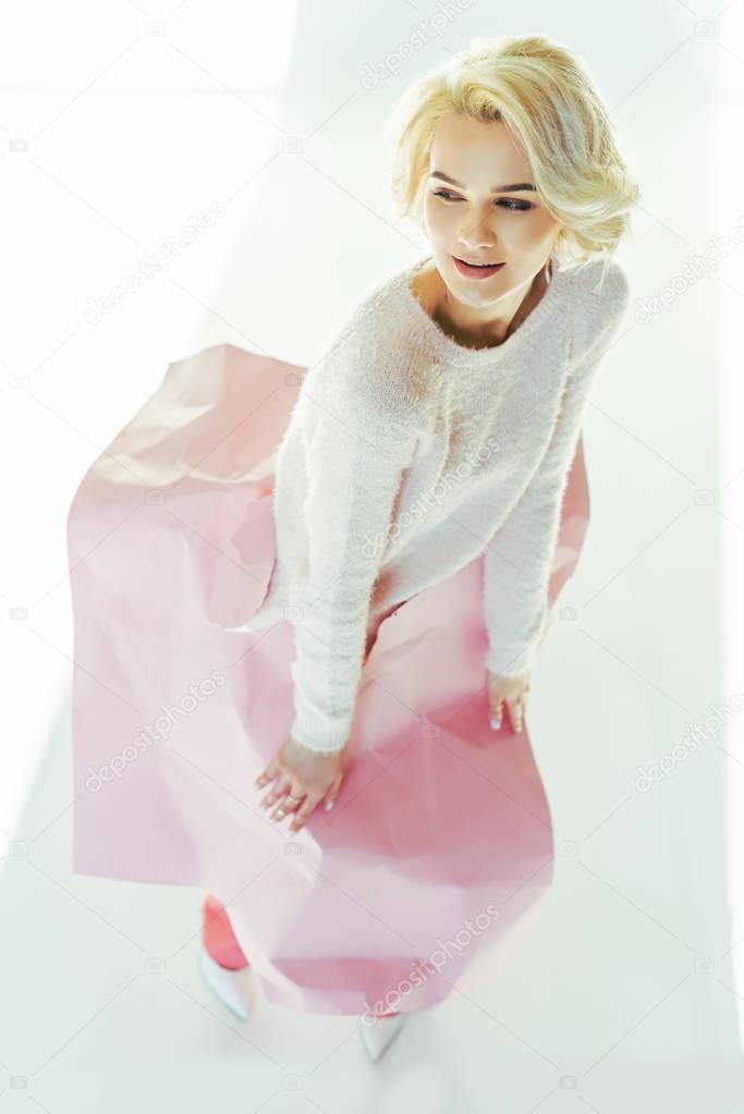 high angle view of beautiful young woman posing with pink crumpled paper on waist and looking away in studio