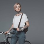 Bella ragazza tatuata con zaino in piedi con la bicicletta e guardando lontano isolato sul grigio