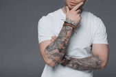 cropped shot of stylish young tattooed man in white t-shirt standing with hand on chin isolated on grey
