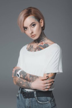 portrait of beautiful girl with tattoos standing with crossed arms and looking at camera isolated on grey clipart