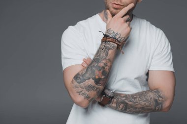 cropped shot of stylish young tattooed man in white t-shirt standing with hand on chin isolated on grey clipart