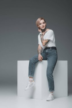 beautiful young woman with tattoos sitting on white cube and looking at camera on grey clipart