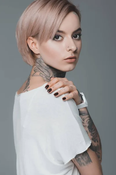 portrait of beautiful tattooed girl in white t-shirt looking at camera isolated on grey