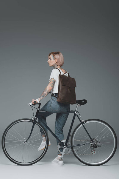 stylish young tattooed woman with backpack sitting on bike and looking away on grey