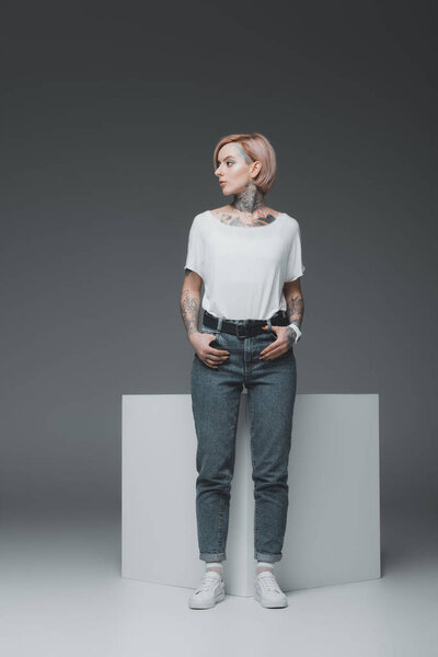 stylish tattooed girl standing with hands in pockets and looking away on grey