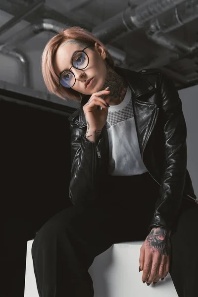 Stylish Pensive Young Woman Eyeglasses Leather Jacket Sitting White Cube — Stock Photo, Image
