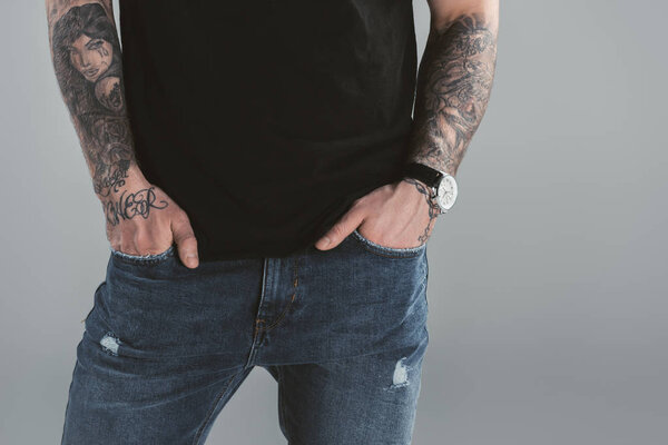 cropped view of tattooed man with watch and hands in pockets, isolated on grey 