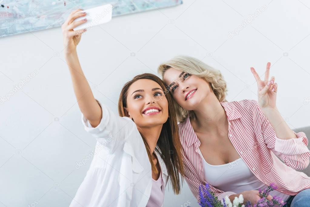 cheerful female friends with victory sign taking selfie on smartphone