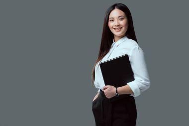 beautiful young asian businesswoman holding folder and smiling at camera isolated on grey clipart