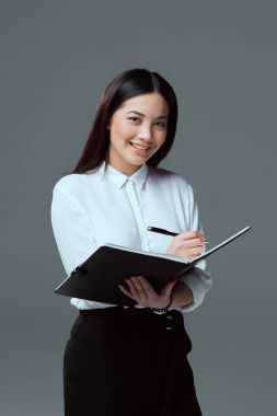 smiling young businesswoman taking notes and looking at camera isolated on grey clipart