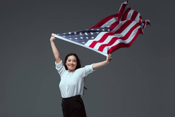 beautiful young woman holding american flag in raised hands and smiling at camera isolated on grey