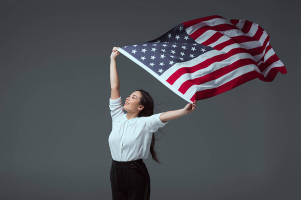 happy young woman holding american flag in raised hands and looking away isolated on grey