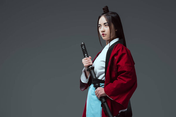 young asian woman in kimono holding katana and looking away isolated on grey