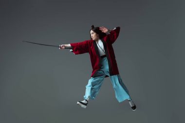 full length view of samurai in kimono fighting with katana in jump isolated on grey clipart
