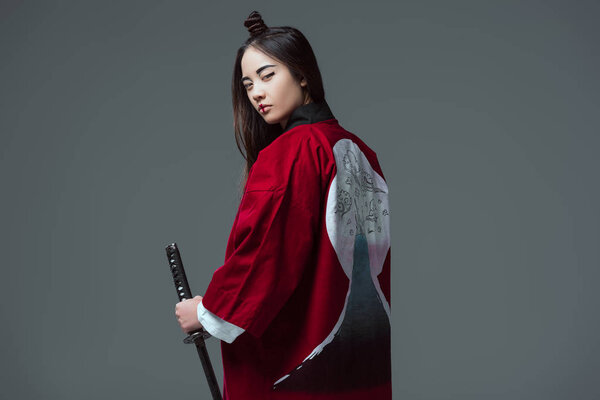 young woman in traditional japanese kimono holding katana and looking at camera isolated on grey 