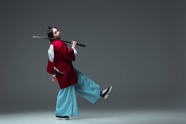 side view of samurai in kimono walking with katana and looking at camera on grey