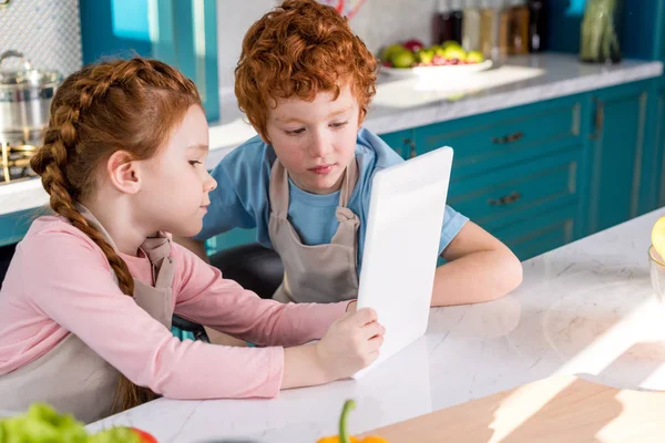 デジタル タブレットを使用して一緒にキッチンで料理中のエプロンでかわいい子供たち — ストック写真