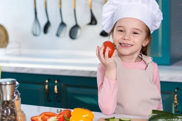 adorable child in chef hat holding tomato and smiling at camera in kitchen