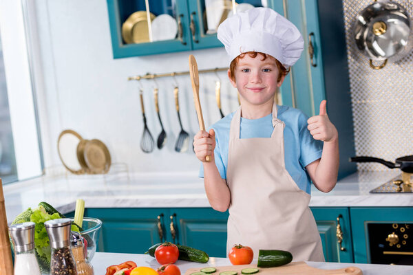 cute little boy in chef hat and apron smiling at camera and showing thumb up in kitchen