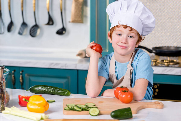 cute little boy in chef hat and apron smiling at camera while cooking in kitchen