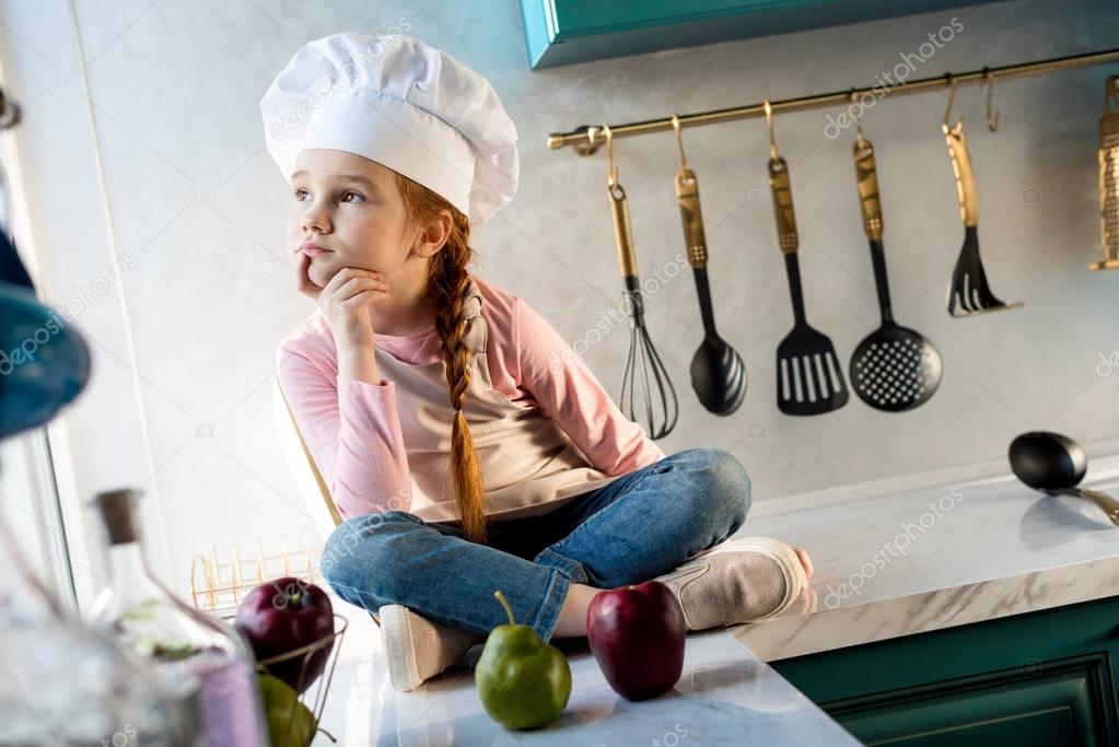 pensive kid in chef hat looking away while sitting in kitchen