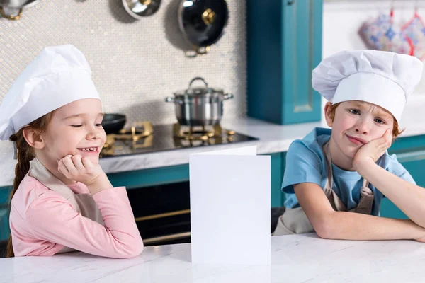 Niedliche Kleine Kinder Kochmützen Sitzen Mit Leerer Karte Der Küche — kostenloses Stockfoto