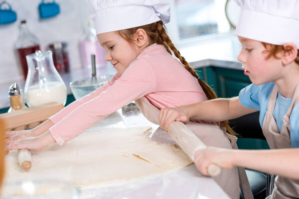 side view of cute little children in chef hats and aprons rolling tough in kitchen
